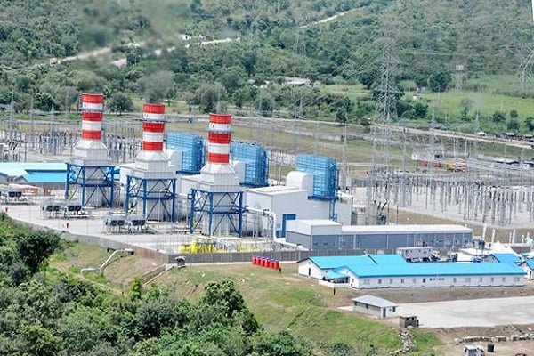 Aerial view of the Geregu gas power plant in Kogi State, with three red-and-white striped exhaust stacks, blue turbine halls and a switchyard with transmission towers, set against green hills.